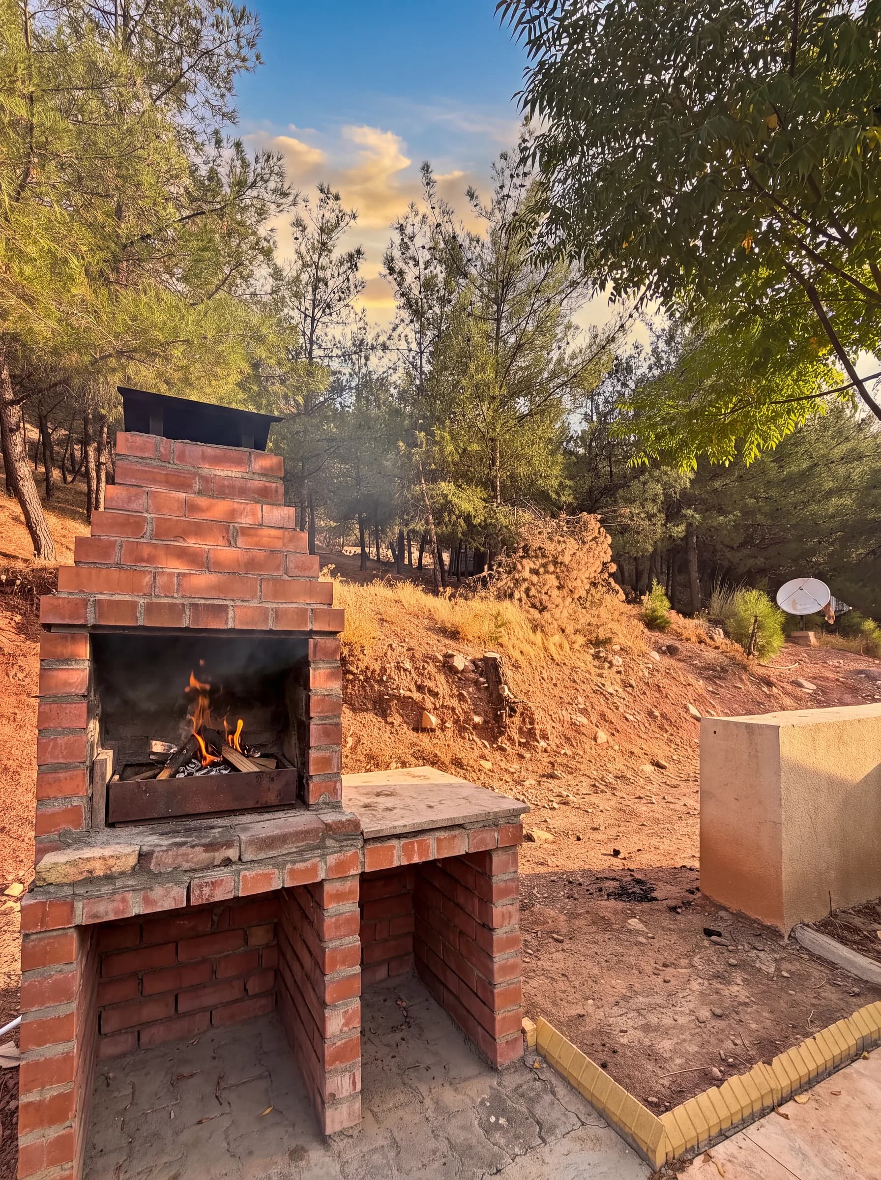 A lit brick barbecue grill on a slope among pine trees