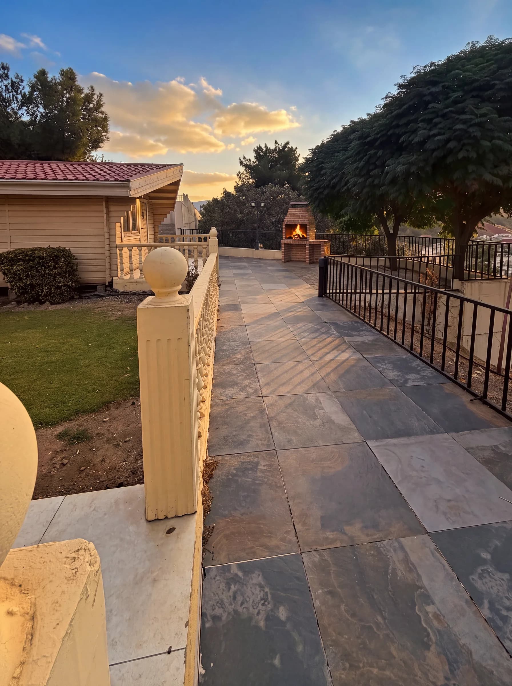 A garden path leading past a cabin to a brick barbecue grill with a wood fire lit, at golden hour
