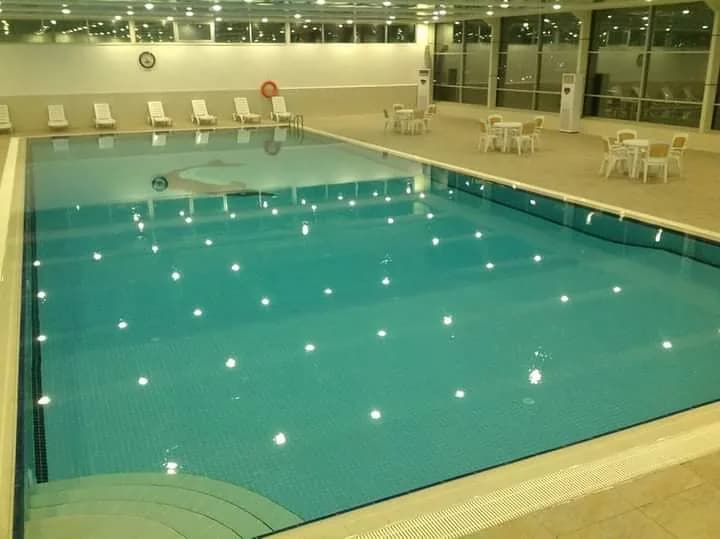 Indoor swimming pool with white loungers and tables beside large windows.
