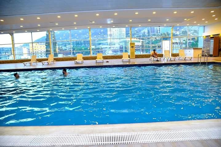 People in and beside an indoor swimming pool, with a town visible through large windows.